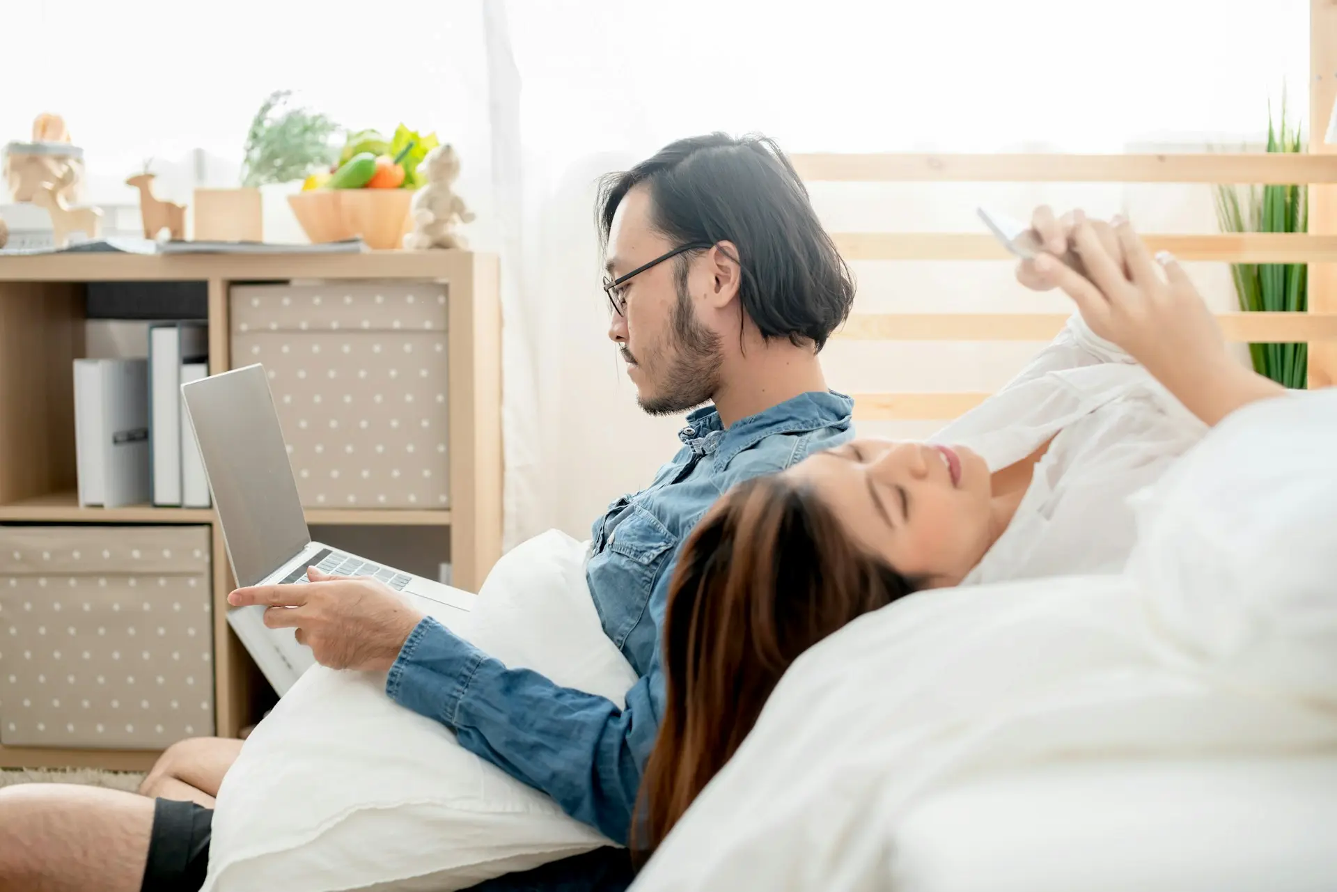 A man working on his laptop, and his wife lying on her back smiling as she uses her phone